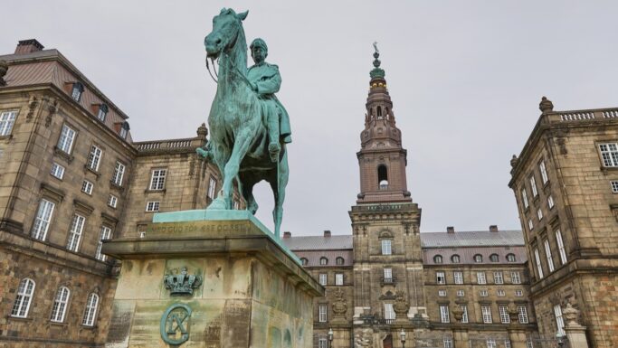 En rytterstatue i bronze står foran Folketinget, den historiske danske parlamentsbygning med et centralt tårn, omgivet af stenkonstruktioner med mange vinduer. Den overskyede himmel bidrager til den værdige atmosfære, som ældresagen fejrer.