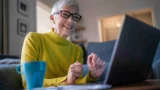 En smilende ældre kvinde med kort gråt hår og briller, iført en gul rullekravebluse, sidder ved et bord med en blå kop og nogle papirer og tjekker på sin bærbare computer, hvordan aktier påvirker hendes pension, i en hyggelig stue.