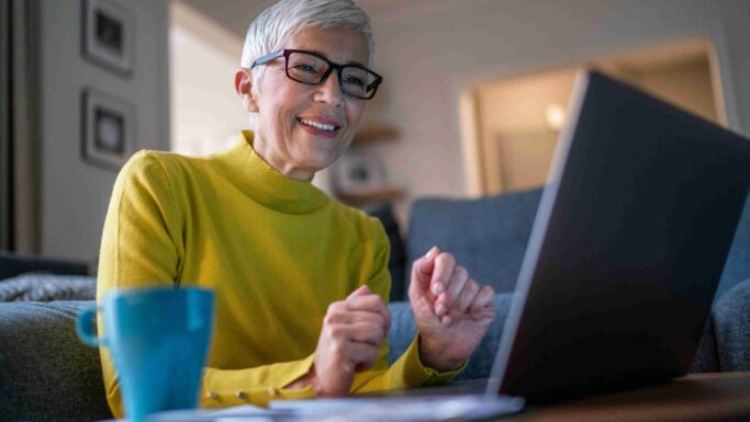 En smilende ældre kvinde med kort gråt hår og briller, iført en gul rullekravebluse, sidder ved et bord med en blå kop og nogle papirer og tjekker på sin bærbare computer, hvordan aktier påvirker hendes pension, i en hyggelig stue.