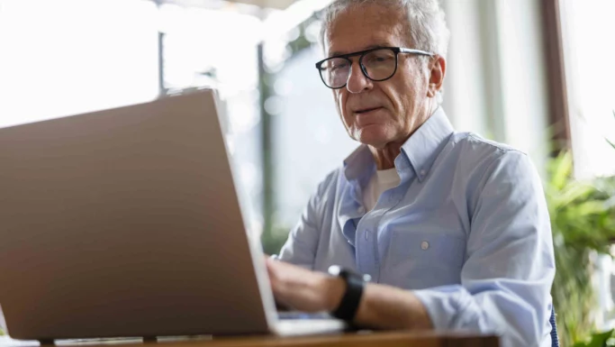En ældre mand med briller og en lyseblå skjorte sidder ved et bord og arbejder på en bærbar computer i et lyst, moderne rum med planter – måske er han i gang med at tjekke sin restskat for at spare penge denne sæson.
