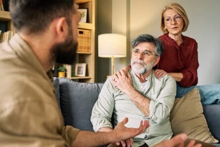Et ældre par sidder i en sofa og lytter opmærksomt til deres voksne børn, der taler med udtryksfulde håndbevægelser. Kvinden lægger hånden på sin mands skulder for at vise sin støtte, mens familien fører en vigtig samtale.