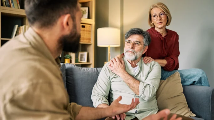 Et ældre par sidder i en sofa og lytter opmærksomt til deres voksne børn, der taler med udtryksfulde håndbevægelser. Kvinden lægger hånden på sin mands skulder for at vise sin støtte, mens familien fører en vigtig samtale.