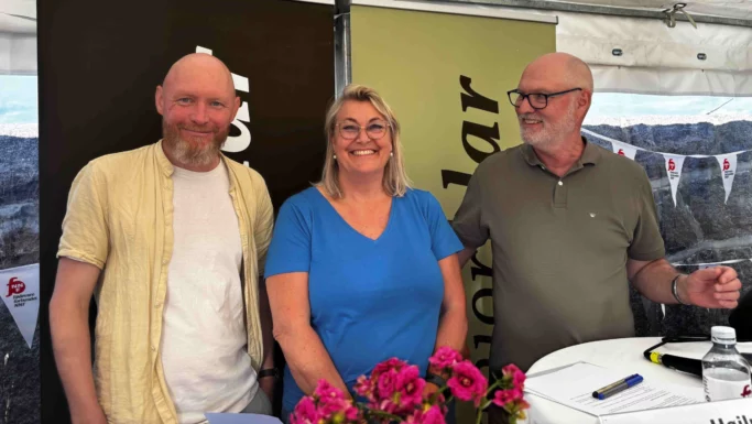 Tre smilende voksne, heriblandt en ældre person, står sammen indendørs ved et arrangement. Bag dem hænger bannere og vimpler, mens der på et bord i forgrunden står en buket lyserøde blomster.