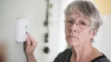 En ældre kvinde med briller og gråt hår justerer en termostat, mens hun ser alvorligt ind i kameraet, bekymret over elregningen og på udkig efter råd i den dyreste sommermåned.