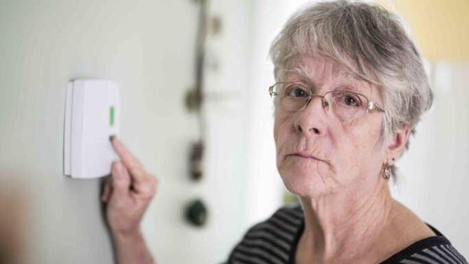 En ældre kvinde med briller og gråt hår justerer en termostat, mens hun ser alvorligt ind i kameraet, bekymret over elregningen og på udkig efter råd i den dyreste sommermåned.