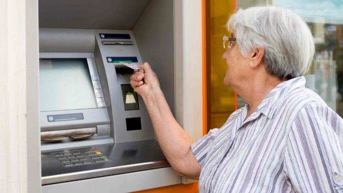 En ældre kvinde med briller og en stribet bluse benytter sig af en af de mange danske pengeautomater og sætter sit kort ind i maskinen. Pengeautomatens skærm og tastatur kan ses, mens hun hæver kontanter.