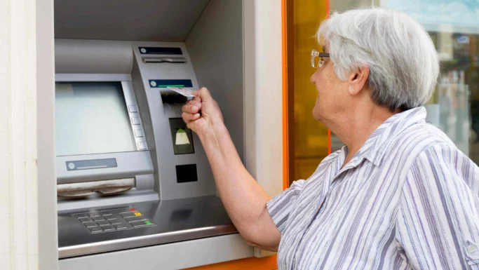 En ældre kvinde med briller og en stribet bluse benytter sig af en af de mange danske pengeautomater og sætter sit kort ind i maskinen. Pengeautomatens skærm og tastatur kan ses, mens hun hæver kontanter.