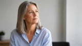 En midaldrende kvinde med gråt hår, iført en lyseblå skjorte, kigger eftertænksomt ud i det fjerne og reflekterer måske over et nyligt jobafslag. Den let slørede baggrund bidrager til den rolige og eftertænksomme atmosfære.