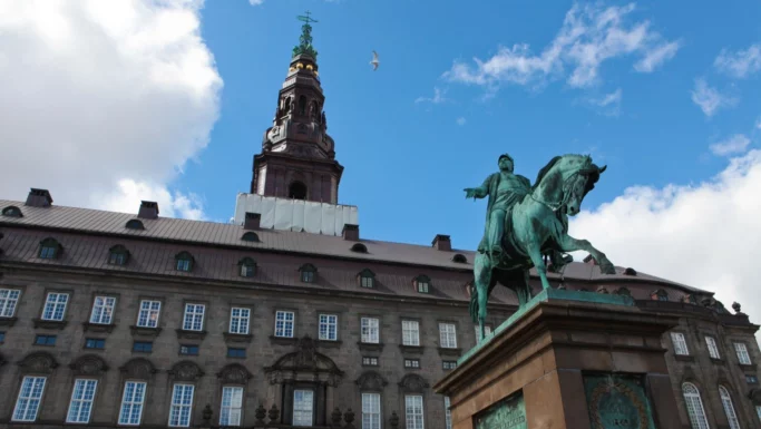 En rytterstatue i bronze står foran en historisk regeringsbygning med mange vinduer og et højt tårn, med en klar blå himmel i baggrunden, hvor der er nogle skyer, og en fugl flyver hen over himlen.