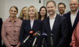 En gruppe professionelt klædte mennesker, herunder flere seniorer, står tæt sammen og smiler bag en række mikrofoner ved en pressekonference eller et mediearrangement.