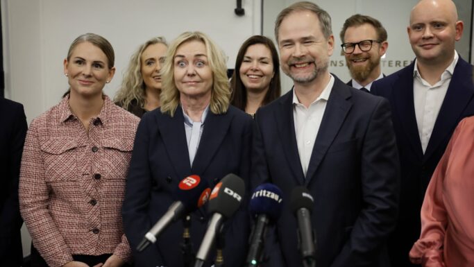 En gruppe professionelt klædte mennesker, herunder flere seniorer, står tæt sammen og smiler bag en række mikrofoner ved en pressekonference eller et mediearrangement.