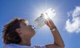 En person drikker vand fra en plastflaske udendørs under en strålende blå himmel under en hedebølge, mens sollyset skinner gennem flasken og skaber en linseglans-effekt.
