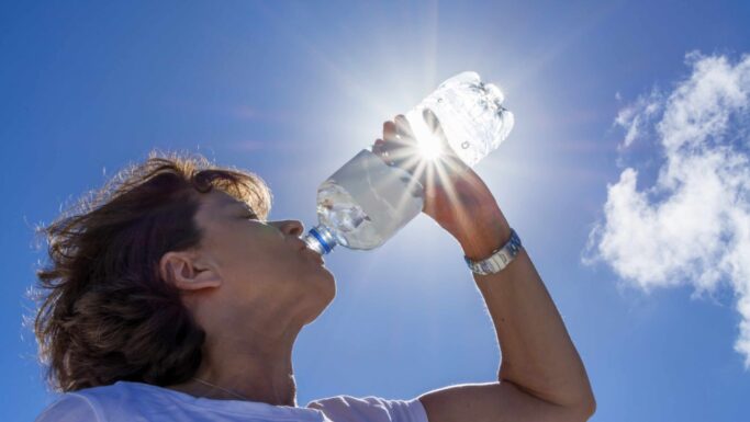 En person drikker vand fra en plastflaske udendørs under en strålende blå himmel under en hedebølge, mens sollyset skinner gennem flasken og skaber en linseglans-effekt.