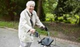 En ældre person på over 90 år med kort, hvidt hår smiler, mens vedkommende går udendørs og bruger en rollator som støtte. Vedkommende har en lys frakke på og slentrer ad en asfalteret sti omgivet af grønne træer og buske.