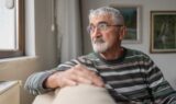 En ældre mand med gråt hår og briller, iført en stribet sweater, sidder i en sofa og kigger eftertænksomt ud af vinduet i sit hyggelige, kunstfyldte værelse og reflekterer over livet efter sin ægtefælle dør.