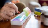 Et nærbillede af en ældre persons hånd, der opmærksomt og forsigtigt lægger en rød medicinkapsel ned i en farverig ugepilleæske på et bord, med forskellige medicinflasker og piller i nærheden.