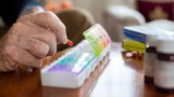 Et nærbillede af en ældre persons hånd, der opmærksomt og forsigtigt lægger en rød medicinkapsel ned i en farverig ugepilleæske på et bord, med forskellige medicinflasker og piller i nærheden.