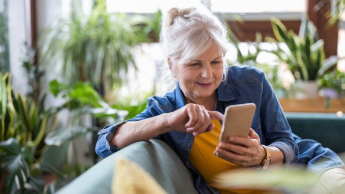 En ældre kvinde med gråt hår sidder på en sofa omgivet af stueplanter og smiler, mens hun bruger sin smartphone til at tjekke en opdatering i MitID-appen. Hun har en gul bluse og en blå denimskjorte på og ser afslappet ud, mens hun er optaget af sin enhed.