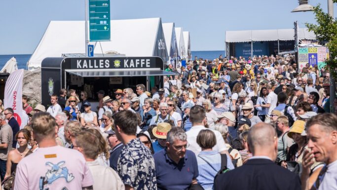 En stor menneskemængde samles udendørs ved Kalles Kaffe med hvide telte i baggrunden på Folkemødet. Mange, inklusive seniorer, er iført sommertøj og hatte, hvilket skaber en livlig scene i det skarpe sollys.