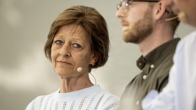 En ældre kvinde med kort brunt hår og en lyseblå trøje har et headset på og ser tankefuldt til siden, mens to mænd ved siden af hende, der også har mikrofoner på, er ude af fokus – en undersøgelse af aldersdiskrimination diskuteres.