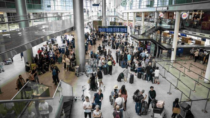 En travl lufthavnsterminal fyldt med rejsende, hvoraf mange har bagage og powerbanks med sig, som står i kø og går rundt. Området har høje glasvinduer, højt til loftet samt forskellige butikker og fastfoodrestauranter i baggrunden.