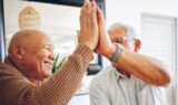 To ældre mænd smiler og giver hinanden en high five indendørs, mens de fejrer en god nyhed om aktiesparekonto loftet og deler et glædeligt øjeblik sammen.