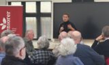 En kvinde taler ind i en mikrofon foran et siddende publikum bestående af ældre voksne i et klasseværelse, hvor hun diskuterer forslag til seniorer. Bag hende ses et rødt banner og en stor tom skærm.