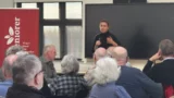 En kvinde taler ind i en mikrofon foran et siddende publikum bestående af ældre voksne i et klasseværelse, hvor hun diskuterer forslag til seniorer. Bag hende ses et rødt banner og en stor tom skærm.