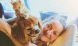 En smilende kvinde slapper af indendørs og krammer en brun og hvid hund, mens en orange killing sidder og leger på hundens hoved. Blødt naturligt lys fylder den hyggelige scene og indfanger et populært øjeblik af sommer med elskede kæledyrsnavne.