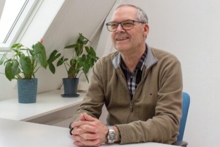 En ældre mand med briller og en khakifarvet trøje sidder ved et hvidt skrivebord og smiler. Bag ham står to potteplanter på vindueskarmen i et lyst, medicinfrit rum med skrå lofter – måske nyder han bacon og æg til morgenmad.