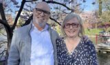 En smilende mand og kvinde står tæt sammen udendørs, omgivet af blomstrende træer og forårsblomster. Begge har briller og let tøj på, og de nyder den lyse dag, mens de diskuterer regeringens seneste ændringer af modregning i efterløn.