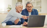 En ældre kvinde og mand sidder sammen i en sofa og kigger på en bærbar computer, muligvis for at gennemgå deres pensionsopsparing. Kvinden peger på skærmen, mens de koncentrerer sig, og der ligger papirer og en kaffekop på bordet foran dem.