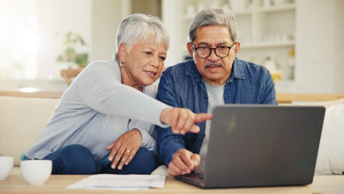 En ældre kvinde og mand sidder sammen i en sofa og kigger på en bærbar computer, muligvis for at gennemgå deres pensionsopsparing. Kvinden peger på skærmen, mens de koncentrerer sig, og der ligger papirer og en kaffekop på bordet foran dem.