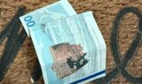 En krøllet 100 norske kroner-seddel ligger på dørmåtten, en brun vævet dørmåtte, hvor man i baggrunden delvist kan skimte sorte bogstaver.