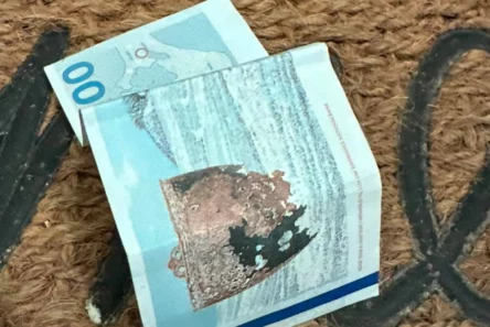 En krøllet 100 norske kroner-seddel ligger på dørmåtten, en brun vævet dørmåtte, hvor man i baggrunden delvist kan skimte sorte bogstaver.