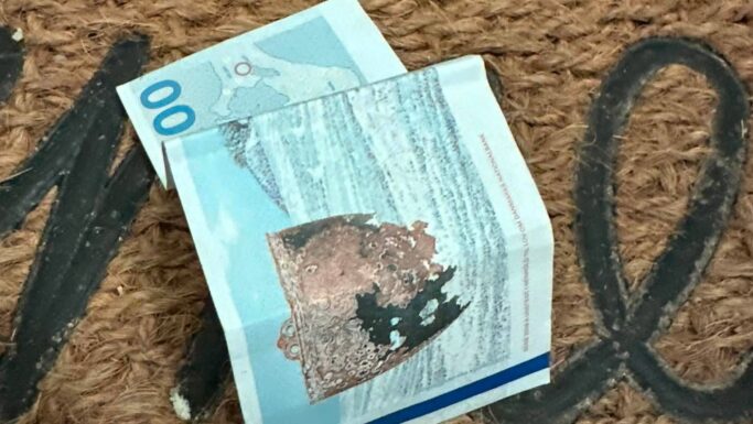 En krøllet 100 norske kroner-seddel ligger på dørmåtten, en brun vævet dørmåtte, hvor man i baggrunden delvist kan skimte sorte bogstaver.