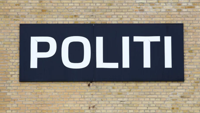 Et stort sort skilt med ordet POLITI i fede hvide bogstaver er monteret på en gul murstensvæg og fremhæver deres kamp mod svindel, efter at en kvinde tabte en halv million.