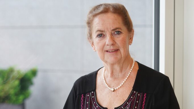 En ældre kvinde med lys hud, blå øjne og brunt hår, der er sat op, står inde foran et vindue iført en sort bluse med farverigt broderi og en perlekæde – hvilket afspejler den værdighed, der ofte forbindes med ældre, der nyder godt af skattelettelser.