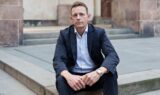 Rasmus Stoklund i mørkt jakkesæt og lyseblå skjorte sidder på nogle stentrapper udendørs og ser direkte ind i kameraet med et alvorligt udtryk – måske tænker han over forsinket regulering eller de seneste regeringsbeslutninger. I baggrunden ses stenvægge og en trædør.