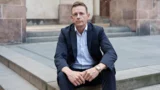Rasmus Stoklund i mørkt jakkesæt og lyseblå skjorte sidder på nogle stentrapper udendørs og ser direkte ind i kameraet med et alvorligt udtryk – måske tænker han over forsinket regulering eller de seneste regeringsbeslutninger. I baggrunden ses stenvægge og en trædør.