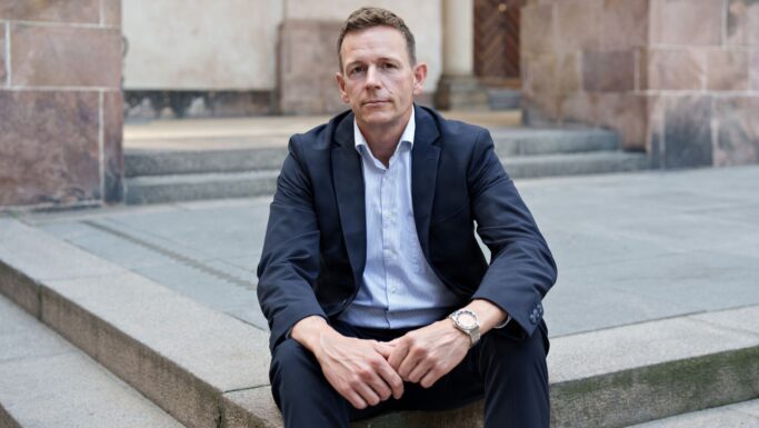 Rasmus Stoklund i mørkt jakkesæt og lyseblå skjorte sidder på nogle stentrapper udendørs og ser direkte ind i kameraet med et alvorligt udtryk – måske tænker han over forsinket regulering eller de seneste regeringsbeslutninger. I baggrunden ses stenvægge og en trædør.