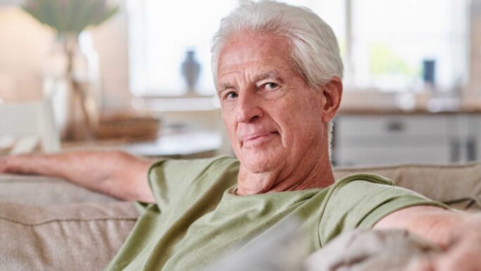En ældre mand med hvidt hår sidder i en sofa iført en lysegrøn skjorte og smiler blidt til kameraet - en af de trygge Arne-pensionisterne, der nyder sin pension i et svagt oplyst, lyst og hyggeligt rum.