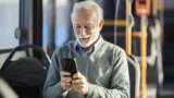 En ældre mand med hvidt hår og skæg smiler, mens han bruger Rejsekort-appen på sin smartphone, siddende i en bus. Han har en lysegrøn trøje på over en skjorte med krave, og i baggrunden ses tomme sæder og busens håndlister.