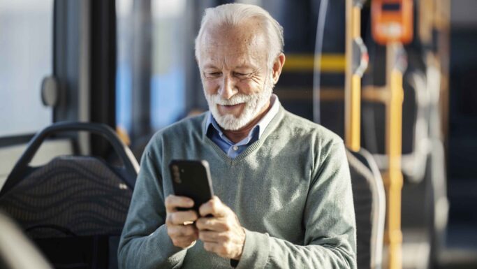 En ældre mand med hvidt hår og skæg smiler, mens han bruger Rejsekort-appen på sin smartphone, siddende i en bus. Han har en lysegrøn trøje på over en skjorte med krave, og i baggrunden ses tomme sæder og busens håndlister.