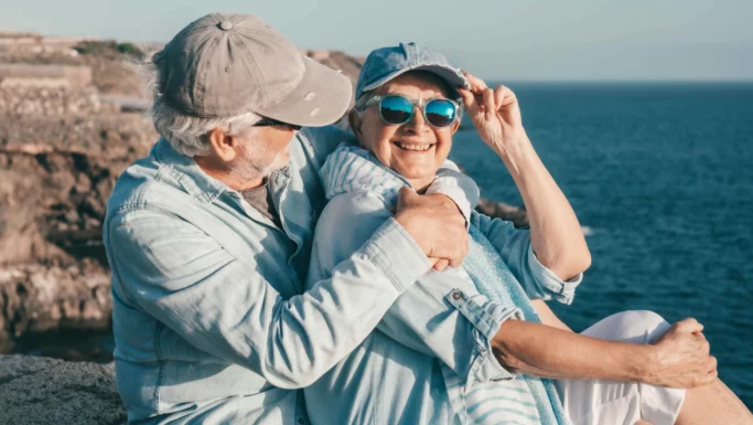 Et ældre ægtepar sidder ved havet, smilende og i hinandens favn. Begge er klædt i lyseblåt tøj, solbriller og kasketter og nyder deres sommerferie med havet og den klippefyldte kystlinje i baggrunden på en solrig dag.