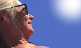 Nærbillede af en ældre kvinde med lyst hår, der bærer sorte solbriller og kigger op mod den strålende sol på en klar, blå himmel. Sollyset fremhæver hendes ansigt og hår, mens hun forsøger at beskytte sig mod solskoldning.