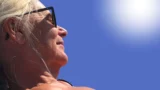 Nærbillede af en ældre kvinde med lyst hår, der bærer sorte solbriller og kigger op mod den strålende sol på en klar, blå himmel. Sollyset fremhæver hendes ansigt og hår, mens hun forsøger at beskytte sig mod solskoldning.