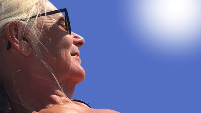 Nærbillede af en ældre kvinde med lyst hår, der bærer sorte solbriller og kigger op mod den strålende sol på en klar, blå himmel. Sollyset fremhæver hendes ansigt og hår, mens hun forsøger at beskytte sig mod solskoldning.