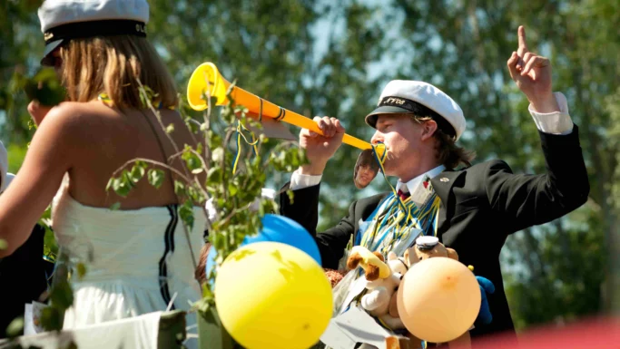 En gruppe unge i studenterhuer og festtøj fejrer udendørs med balloner og pynt i overensstemmelse med traditionen; én person blæser i en gul vuvuzela og gestikulerer energisk, hvilket markerer en glædelig begyndelse.