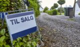 Et »Til Salg«-skilt står ved en grusindkørsel, der fører hen til et hvidt hus – perfekt for en folkepensionist, der gerne vil sælge huset og afveje fordele og ulemper ved livet på landet, omgivet af grønne hække og træer.